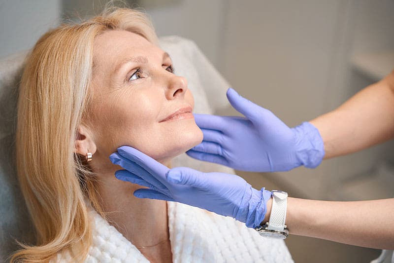 Hands in medical gloves touching happy woman face older woman having a cosmetic procedure